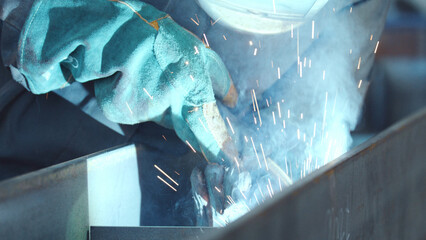 Industrial welder creating a bright blue arc with flying sparks and smoke, wearing helmet and gloves while joining steel components in a workshop, showing skilled metalwork