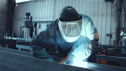 Skilled industrial worker wearing protective gear and safety equipment while joining metal elements with welding sparks, concentrating on the precision task in a professional factory setting