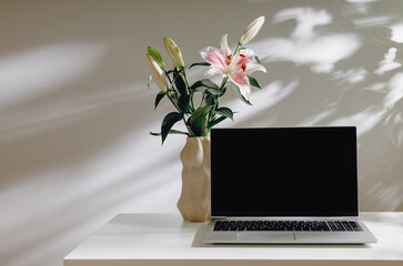 Aesthetic laptop mockup with black screen and stylish vase with gentle lily flower bouquet on white table with beautiful shadows. Laptop for web site promotion, blog, design,home office or branding