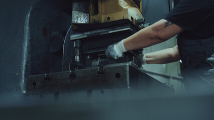 Worker's gloved hands operating a heavy hydraulic press to bend and stamp steel parts in a workshop, showcasing precision metalworking and skilled industrial production processes