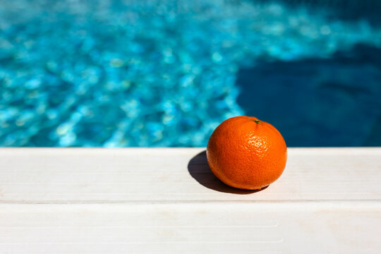 a singular clementine placed at the edge of a pool with bright blue pool water shining in the background