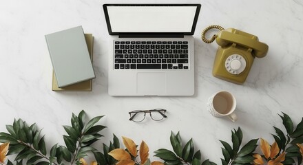 A top-down view of a laptop books an olive green rotary phone eyeglasses and a mug on a white marble surface with green and yellow leafy branches at the bottom