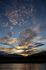 Breathtaking sunset landscape, illuminating sky with vibrant orange and gold clouds. Tranquil water reflects stunning colors, creating peaceful scene above majestic mountain silhouettes