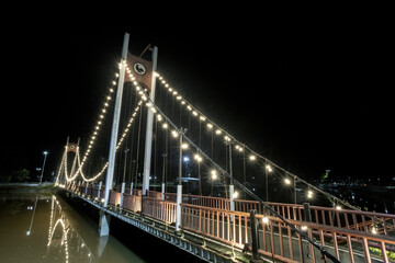 Obraz premium Illuminated suspension bridge shines brightly at night, casting beautiful reflection upon calm water. Its glowing urban architecture creates tranquil scene
