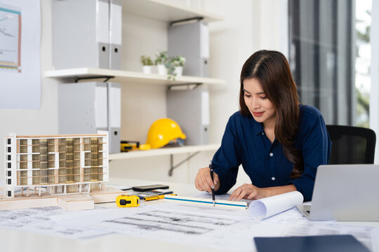 Young female engineer designer or contractor working on drawing plans and making measurements on blueprints of post tension slab plan by triangular scale at workplace, copy space - Powered by Adobe
