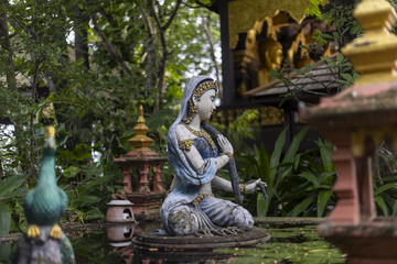 Fototapeta premium Stone sculpture depicting serene female deity, ornate traditional Asian style. Figure sits gracefully within lush spiritual garden. Ancient statue evokes deep sense of peaceful tranquility