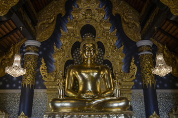 Fototapeta premium Serene golden Buddha statue sits peacefully within richly decorated temple interior, surrounded by intricate gold carvings and dark blue walls. sacred space exudes profound tranquility