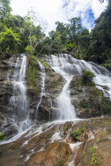 Fototapeta premium Serene waterfall cascades gracefully over mossy rocks lush tropical forest. Clear water flows smoothly, creating tranquil and peaceful natural scene bright sky, highlighting pure beauty of nature