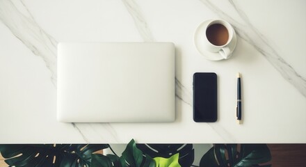 Overhead view of a minimalist workspace on white marble featuring a laptop smartphone pen and a cup of beverage next to green foliage