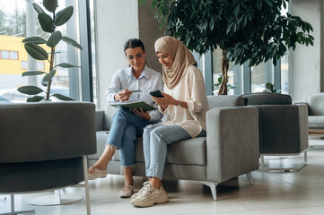 Fototapeta premium In traditional Muslim clothing. Two women are indoors in the modern hall of building at daytime