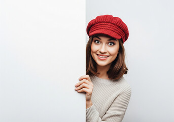 A smiling woman in a red cap peeks from behind a white board, looking at the viewer.
