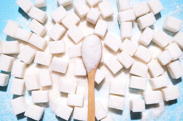 Scattered cubes of white refined sugar and a wooden spoon with granulated sugar on blue background top view flat lay. Concept of eating sweets, health and diabetes issue, sugar addiction