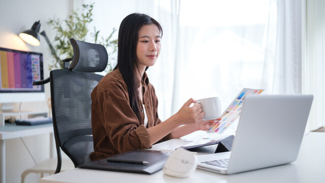 Young creative woman holding a coffee mug and reviewing a color palette chart in a bright, modern workspace. - Powered by Adobe
