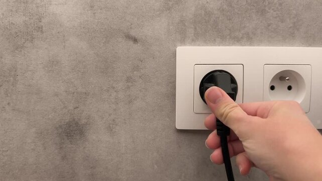 Hand inserting electrical plug into wall socket, demonstrating the connection process with textured gray wall background and dual outlets
