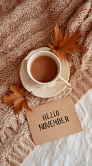 Cup of coffee on knitted blanket with autumn leaves and hello november card in cozy setting