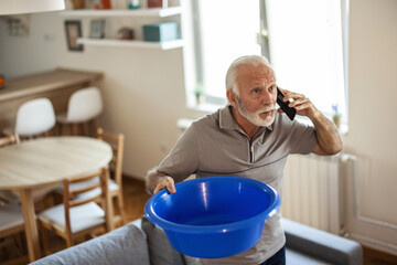 Worried home owner dealing with water leak in the kitchen . A senior Caucasian man collecting water from a leaking rook in his apartment.