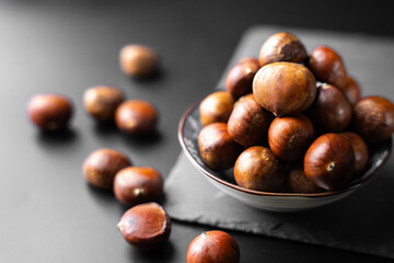 Fresh raw chestnuts in a bowl on black background, healthy organic autumn food, natural seasonal nut rich in flavor and nutrition, perfect ingredient for cooking and roasting.