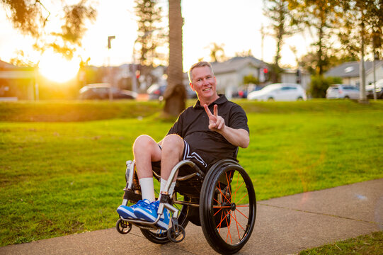 Man in wheelchair flashes a peace sign while sitting on footpath in park during sunset
