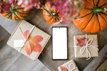 Top-down of smartphone with blank white screen, pumpkins and gift boxes on textured background. Perfect for showcasing app interfaces and e-commerce products during the fall holiday season