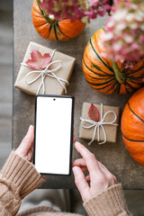 Top view of woman holds smartphone with blank white screen, surrounded by pumpkins, gifts wrapped in craft paper and autumn leaves. Ideal for displaying app concepts or holiday promotions