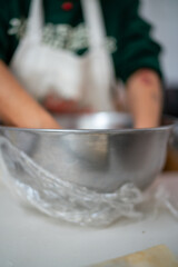 Hands Kneading Dough in a Metal Bowl Covered with Plastic Wrap on a White Countertop