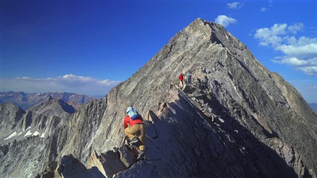 People hiking Knifes Edge North East Ridge trail Capitol Peak Wilderness Trail scrambling rock climbing Rocky Mountains Colorado 14er mountainside boulders summer blue sky morning sunny rugged terrain