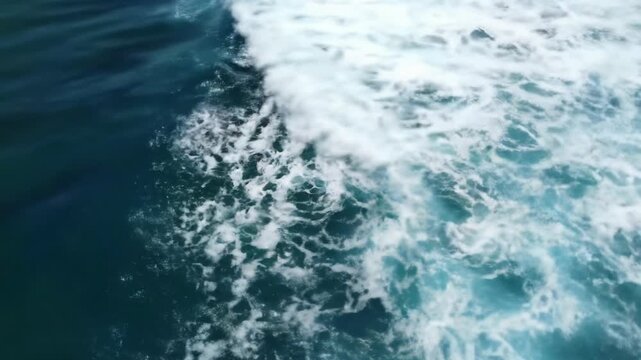 An aerial top-down video of the vast sapphire blue and turquoise ocean, powerful white waves sculpting intricate foam patterns under glistening sunlight with subtle lens glints. Raw power of nature