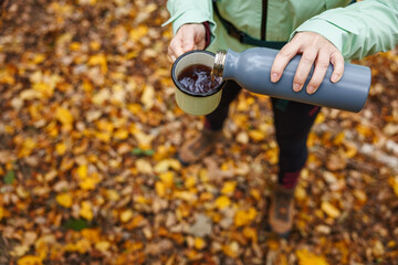 Woman hiker pours tea from thermos into travel mug. Refreshment with hot drink during autumn hiking in forest