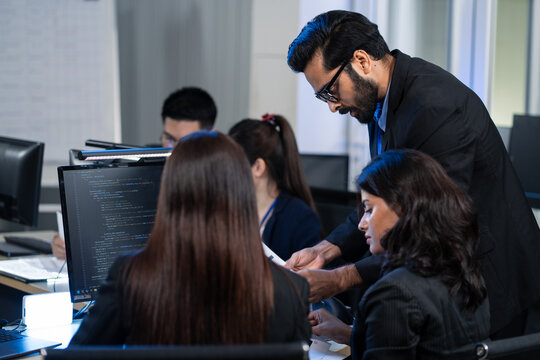 A focused Indian IT manager provides mentorship to diverse team while reviewing computer code on a desktop monitor. Demonstrating strong teamwork and corporate strategy, technical training - Powered by Adobe