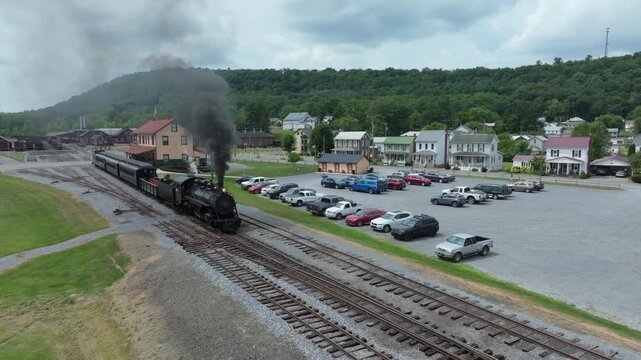 A historic steam train puffs billowing smoke as it moves through a charming town. View the picturesque landscape, vintage buildings, and parked cars on a warm summer afternoon.