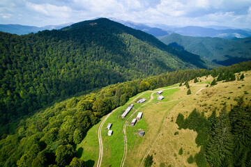 Aerial shot of scenic rural area in Ukraine, featuring series of rustic buildings along winding dirt road. Forested hills provide stunning backdrop, extending into horizon under partly cloudy sky.