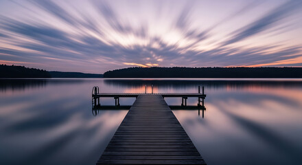 Obraz premium Wooden dock extends onto a calm lake under a colorful sunset with a long exposure effect on the clouds.
