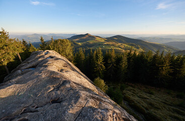 Stunning view from top of rocky outcrop overlooking Pysanyi Kamin in Carpathians, Ukraine. Background reveals rolling green hills, dense forests, and distant mountain ranges under clear blue sky.