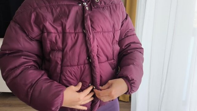 Individual in a violet puffer jacket is fastening buttons, showcasing the action of dressing and preparing for colder weather in a cozy indoor setting