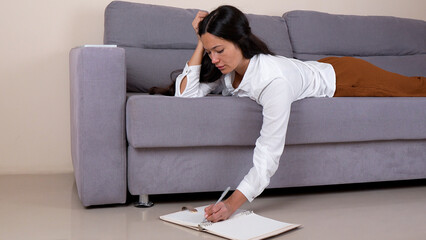 The woman is lying on a sofa and writing in a notebook on the floor