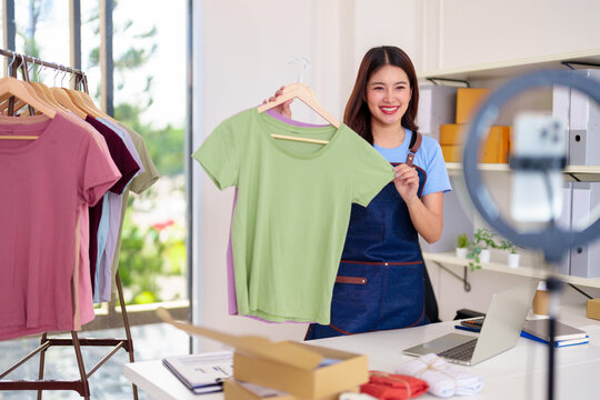Asian adult woman entrepreneur selling fashionable apparel online live streaming from home studio with laptop and clothing rack showcasing merchandise