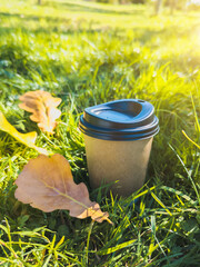 Eco cardboard coffee cup with yellow autumn leaves on green grass. Autumn mood, hot drinks in cold weather. Vertical photo.