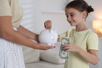 Mother and her daughter putting pocket money into piggy bank at home, closeup