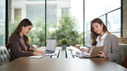 Two professional women collaborating in a well-lit office with laptops and documents.