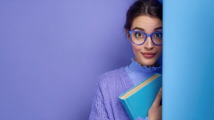 Young Caucasian woman with brown hair and glasses, holding a blue book, standing against a purple background. She has a playful expression.