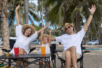 Joyful family celebrating beach vacation with hands up. Parents and child wearing summer outfits and straw hats, sitting under palm trees with colorful drinks on a tropical sunny day.