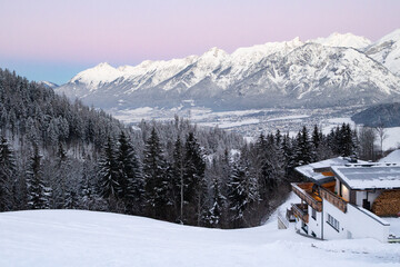 Fototapeta premium Snow-covered mountains rise majestically in the background, with a cozy wooden chalet nestled among evergreen trees, creating a serene winter landscape at dusk