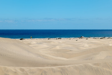View through the sand dunes of Maspalomas towards the Atlantic Ocean. Undulating desert landscape meets beach and deep blue sea under a clear sky.