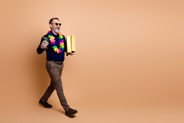 Cheerful mature man carrying folders and a drink in tropical attire walking against beige background