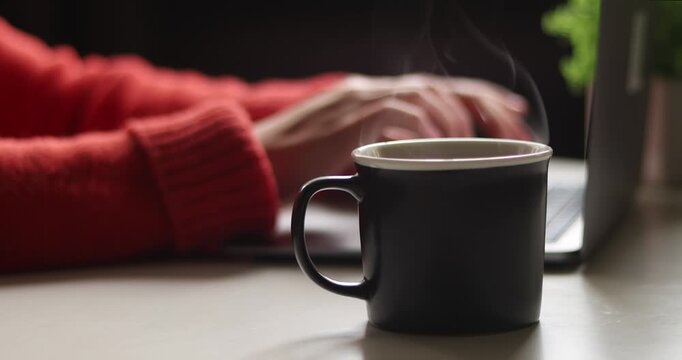 Close-up of laptop on table with hand in red sweater, taking a black  coffee cup onto table surface abd typing on keyboard, blurred background
