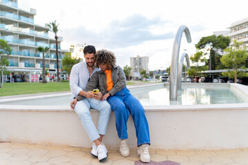 Young couple sharing smartphone, enjoying urban park fountain