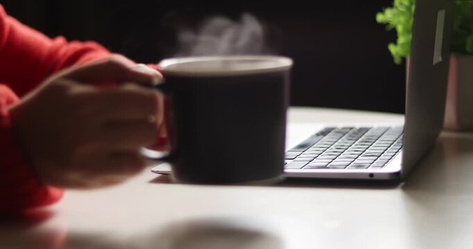 Close-up of laptop on table with hand in red sweater, taking a black  coffee cup onto table surface abd typing on keyboard, blurred background
