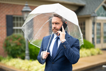 Businessman talks on the phone while standing in the rain. Man in suit with umbrella and makes a mobile phone call. Business man checks his smartphone on a rainy street. Call while rain drops fall.