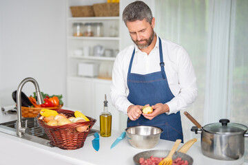 Cook chops vegetables in kitchen. Chef in apron prepares healthy meal in modern kitchen. Cook with vegetables in white kitchen. Cook prepares breakfast with veggies at home kitchen.