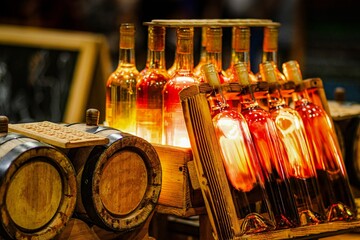 Bottles of amber liquid are displayed on a wooden shelf beside barrels, illuminated by warm light, creating a cozy atmosphere in a rustic setting